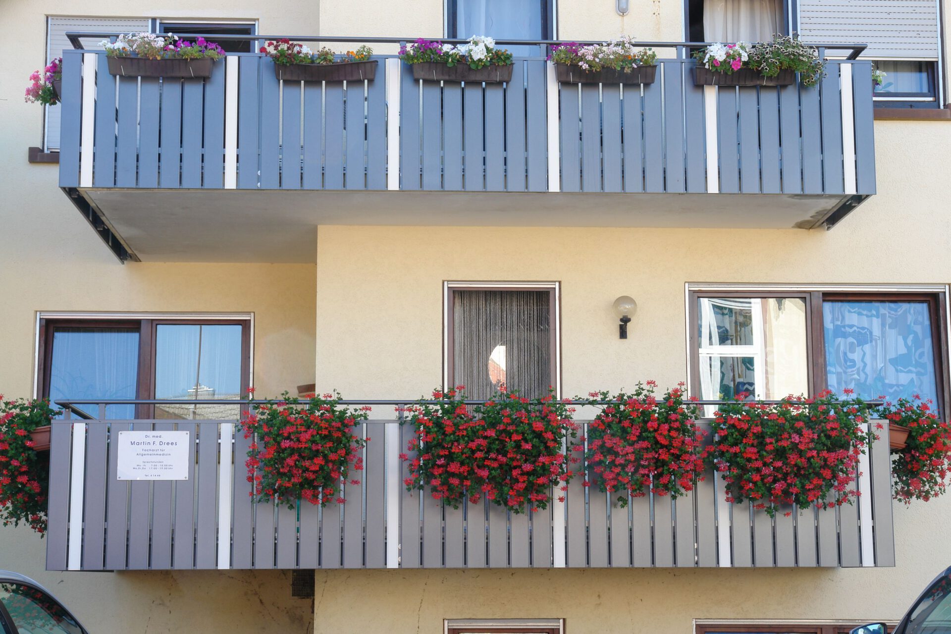 Balkongeländer modern Anthrazit. Gelbes Haus, mit einem Balkon, an dem Blumenkästen hängen. Die Balkonbretter sind aus Aluminium in der Farbe Anthrazit
