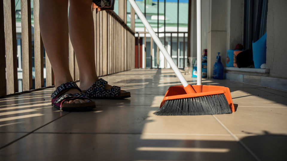 Das Bild zeigt eine Person, die einen Balkon reinigt. Ein Besen und Kehrschaufel sind im Vordergrund sichtbar, während Tageslicht und Schatten ein gepflegtes und ordnungsbewusstes Ambiente betonen.