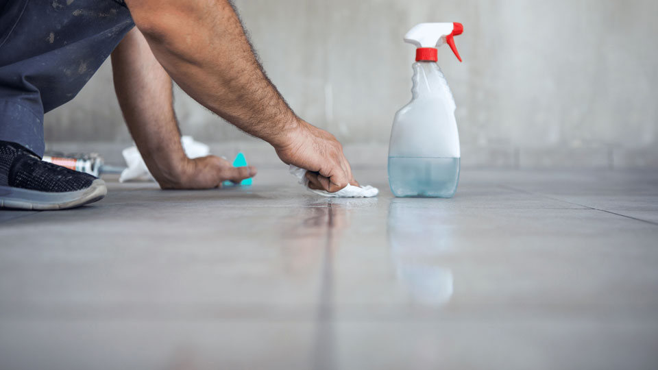 Das Bild zeigt eine Person, die mit einem Tuch den Boden reinigt, während eine Sprühflasche mit Reinigungsmittel daneben steht. Die Szene vermittelt eine gründliche Reinigungssituation.