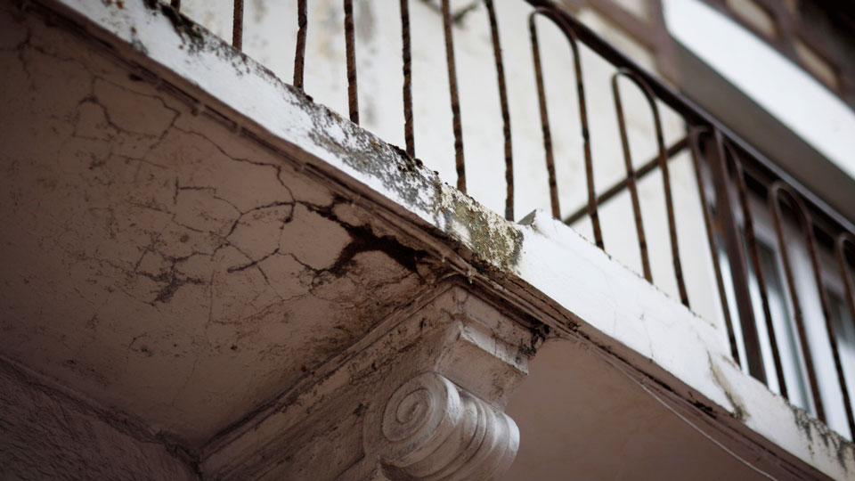 Das Bild zeigt Schäden an einem Balkon, wie Risse im Putz und mögliche Korrosion der Geländerstruktur. Diese Probleme deuten auf Feuchtigkeitseinwirkung und Alterung hin, was eine Sanierung erforderlich macht.