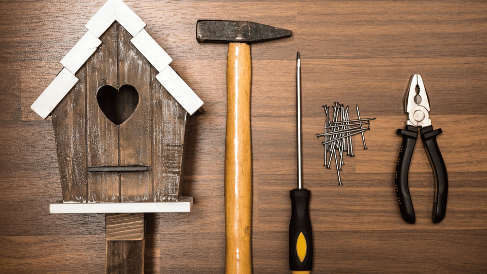 Materialien für den Bau eines Vogelhauses: Ein Holzhaus mit Herz-Ausschnitt, ein Hammer, ein Schraubenzieher, Nägel und eine Zange, auf einem Holztisch arrangiert.