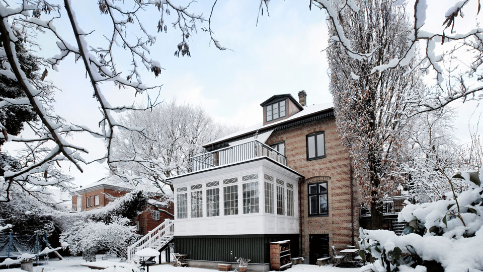  Das Bild zeigt ein großes Backsteinhaus im Winter, umgeben von schneebedeckten Bäumen und Sträuchern. Ein Wintergarten mit vielen Fenstern und einem Balkon darüber hebt sich an der Seite des Hauses hervor. Die kahlen, schneebedeckten Äste der Bäume rahmen das Haus ein und verstärken die ruhige, winterliche Atmosphäre.