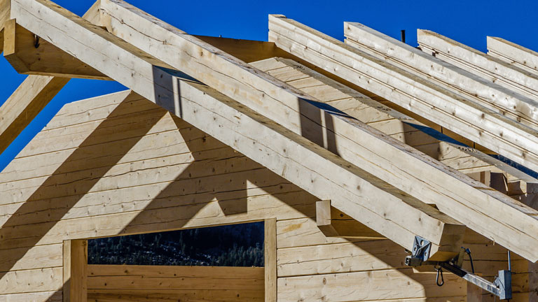 Das Bild zeigt den Rohbau eines Holzhauses mit freiliegenden Balken für das Dach. Die Struktur ist aus hellem Holz gebaut, und die Balken werfen Schatten auf die Wände. Der blaue Himmel im Hintergrund verleiht der Szene eine klare und sonnige Atmosphäre, die den handwerklichen Charakter des Bauprojekts hervorhebt.